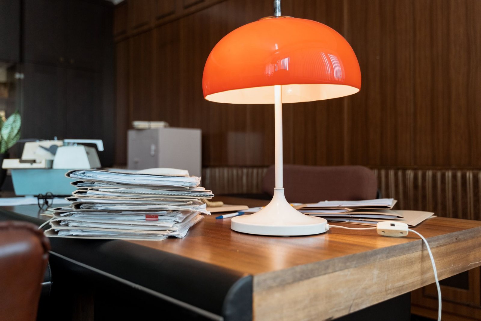 A lit desk lamp at the corner of a home desk with a stack of opened mail squared beside it and a closed folder with a pen resting on top